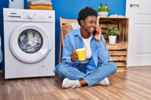 African american woman talking on the smartphone and drinking coffee waiting for washing machine at laundry room